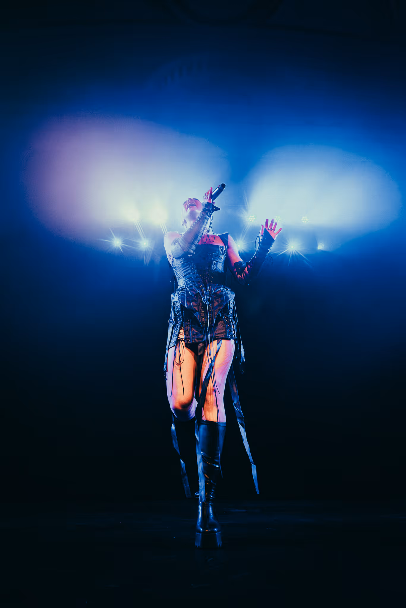 Singer on stage in black outfit and boots under bright blue stage lights with microphone in hand.