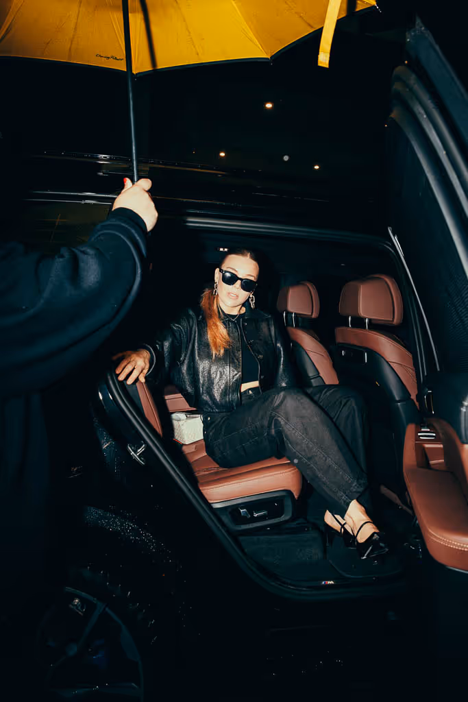 Woman in black outfit and sunglasses sitting in a car's open backseat under a yellow umbrella at night.