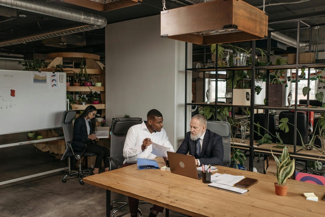 Two professionals having a discussion at a desk in a modern office, while a third person works in the background.