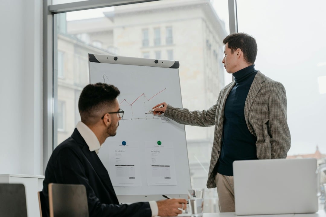 A man presenting data on a whiteboard during a meeting, with another person listening and taking notes. The graph shows trends.