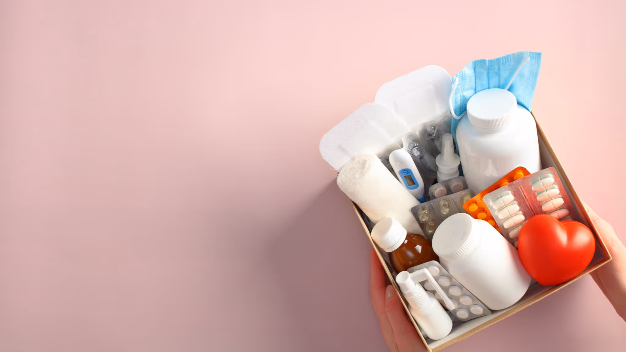 Hand hält einen Karton mit medizinischen Hilfsmitteln wie Pillen, Flaschen, Verband, Fieberthermometer, OP-Maske und rotem Herzmodell.
