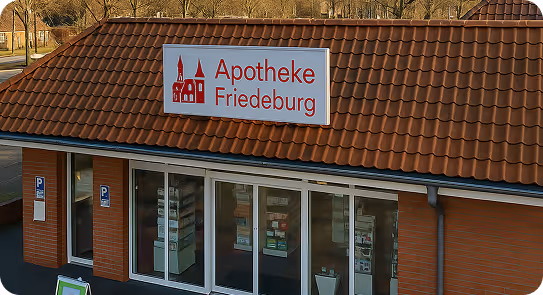 Außenansicht der Apotheke Friedeburg mit rotem Ziegeldach und Schild mit roter Schrift und einem Kirchen-Symbol.