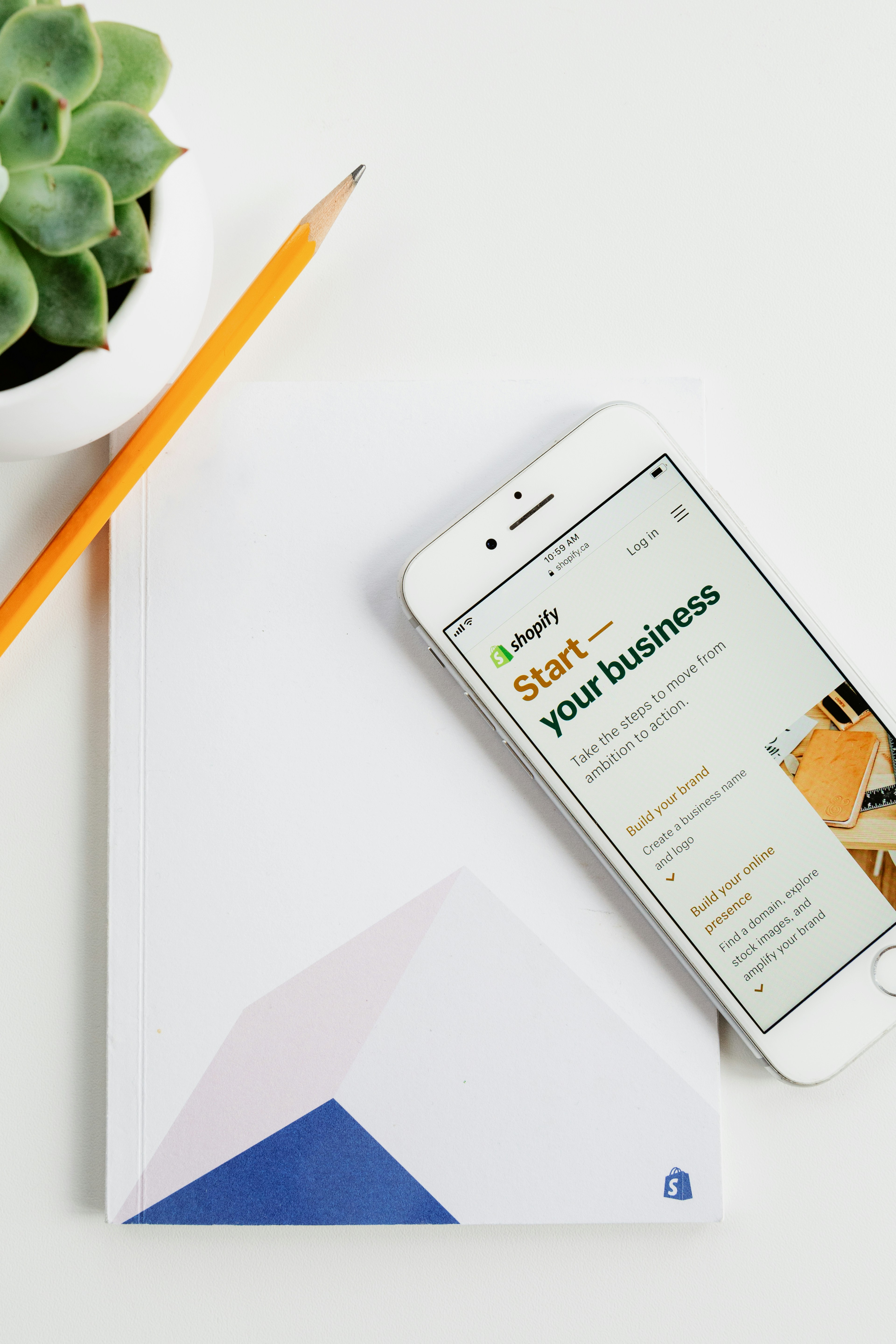 Smartphone showing a Shopify page titled 'Start your business' on a white notebook with a pencil and plant nearby.