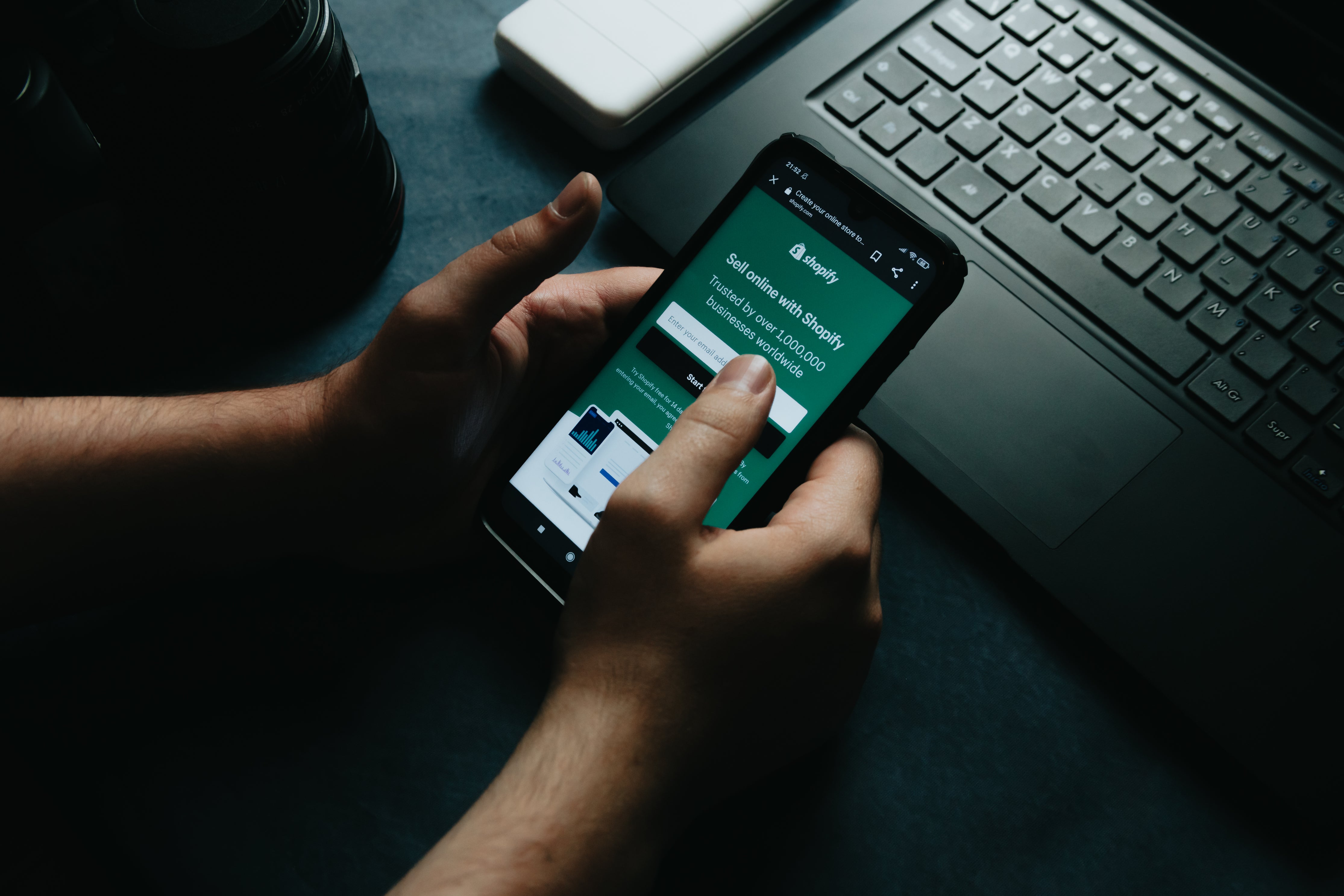 Hands holding a phone with a Shopify screen and a laptop in the background