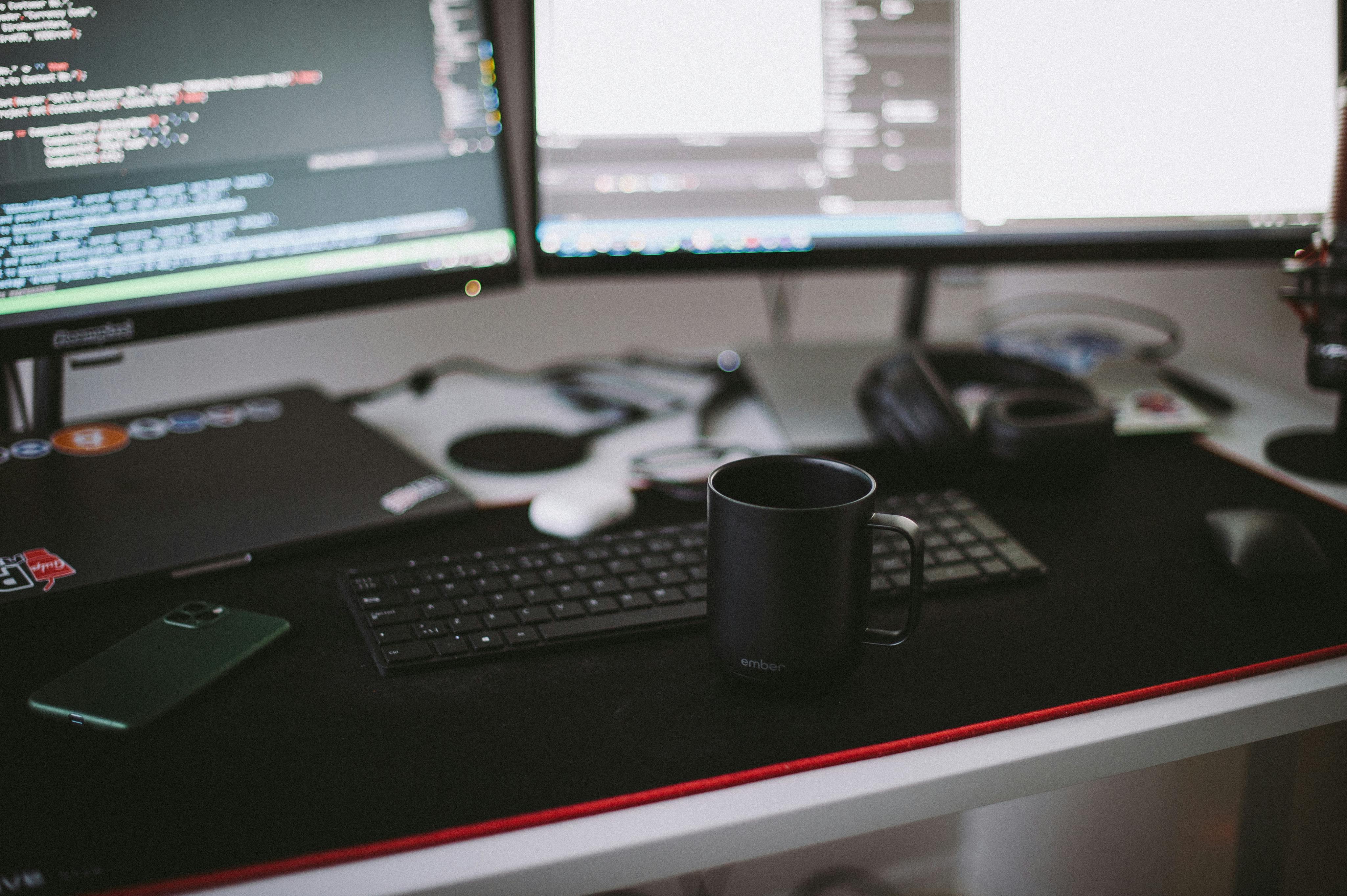 Desk setup with dual monitors showing code, keyboard, black Ember mug, headphones, and smartphone.