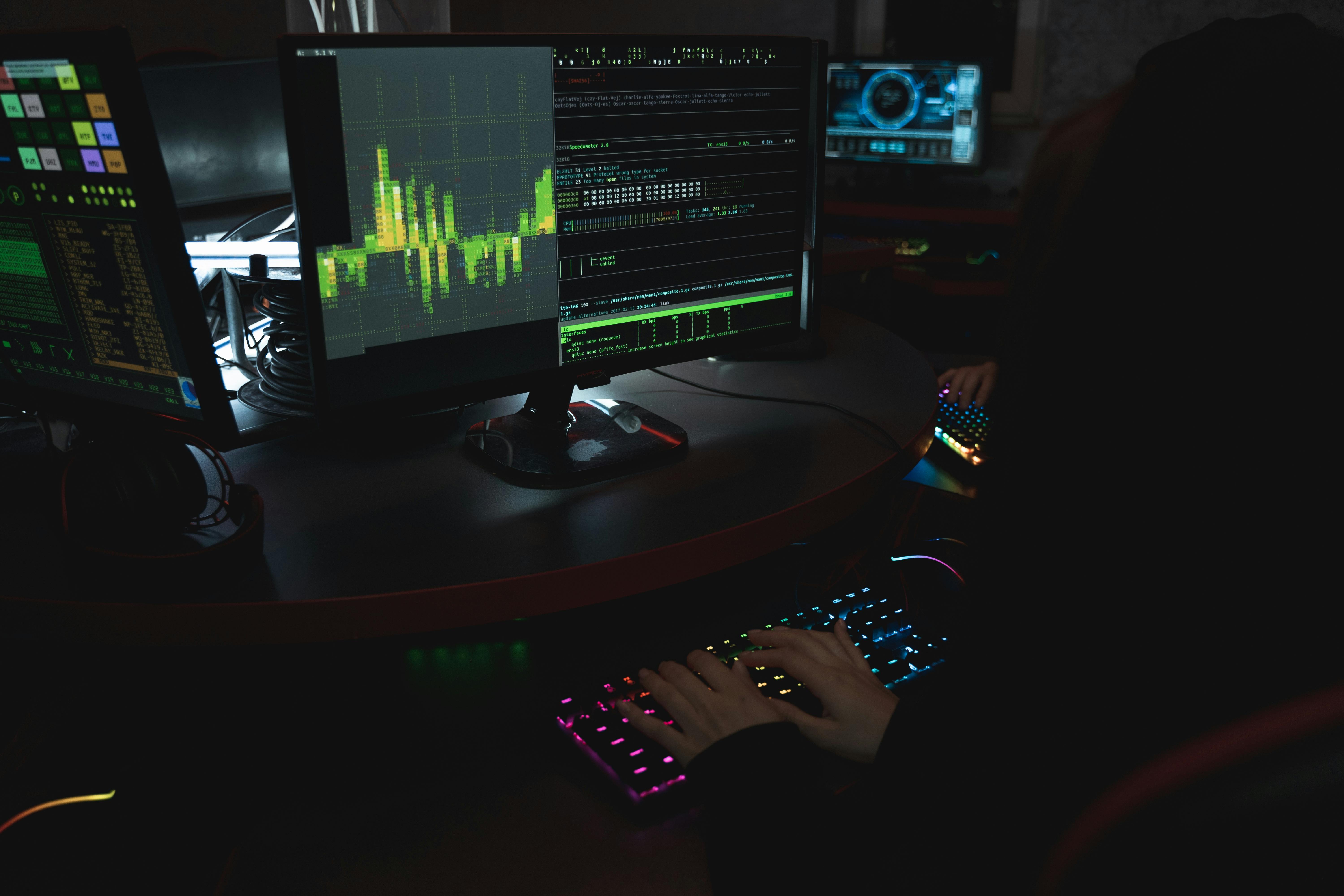 Person typing on a colorful backlit keyboard in a dark room with multiple monitors showing graphs and data.
