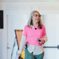 Smiling woman wearing glasses and a pink sweater holding a microphone, standing in a bright room.