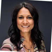 Smiling woman with long dark hair wearing a patterned blouse against a dark background.