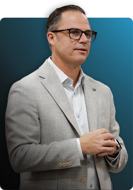 Man with short dark hair and glasses wearing a light gray blazer and white patterned shirt, holding an object with both hands while speaking or presenting.