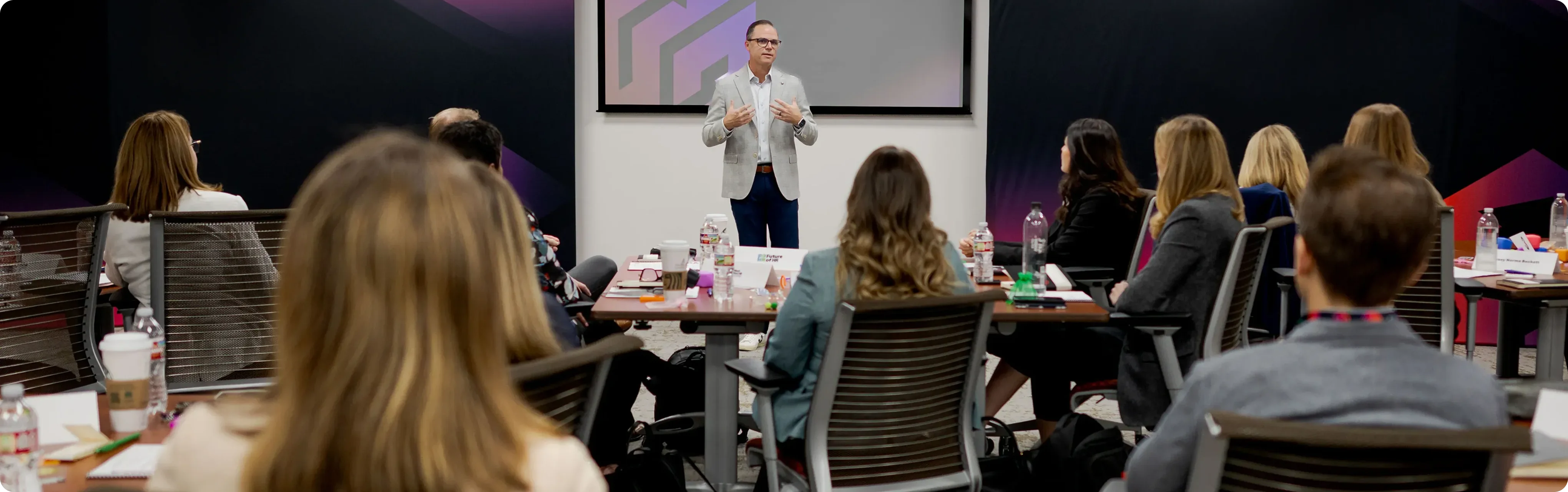 A man in glasses and a light jacket stands and speaks in front of a seated audience in an office or conference room setting.