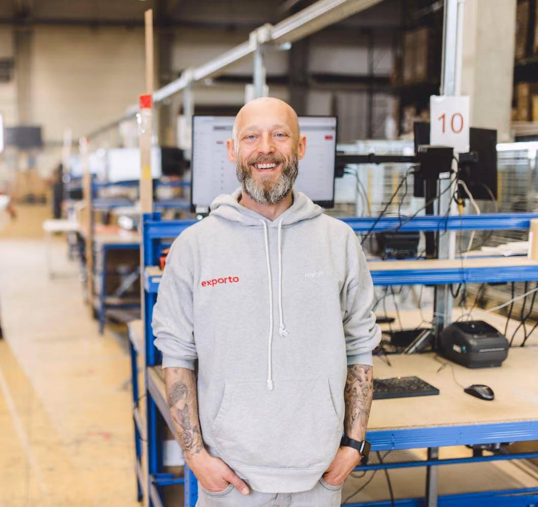 Ein Mann in grauem Pullover steht mit dem Rücken vor einer Workbench und schaut lächeln in die Kamera.