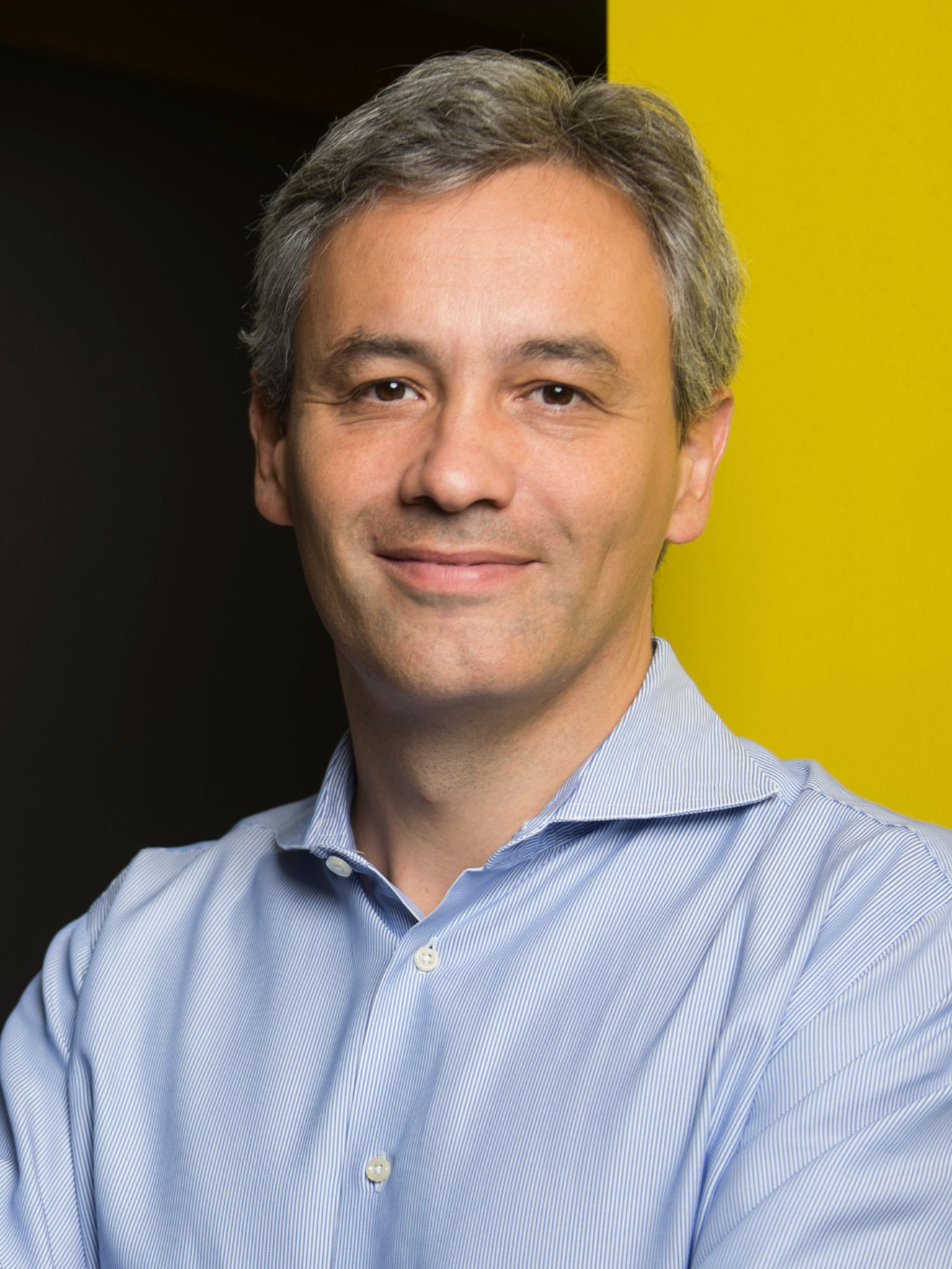 Professional headshot of a middle-aged man in blue shirt against yellow background