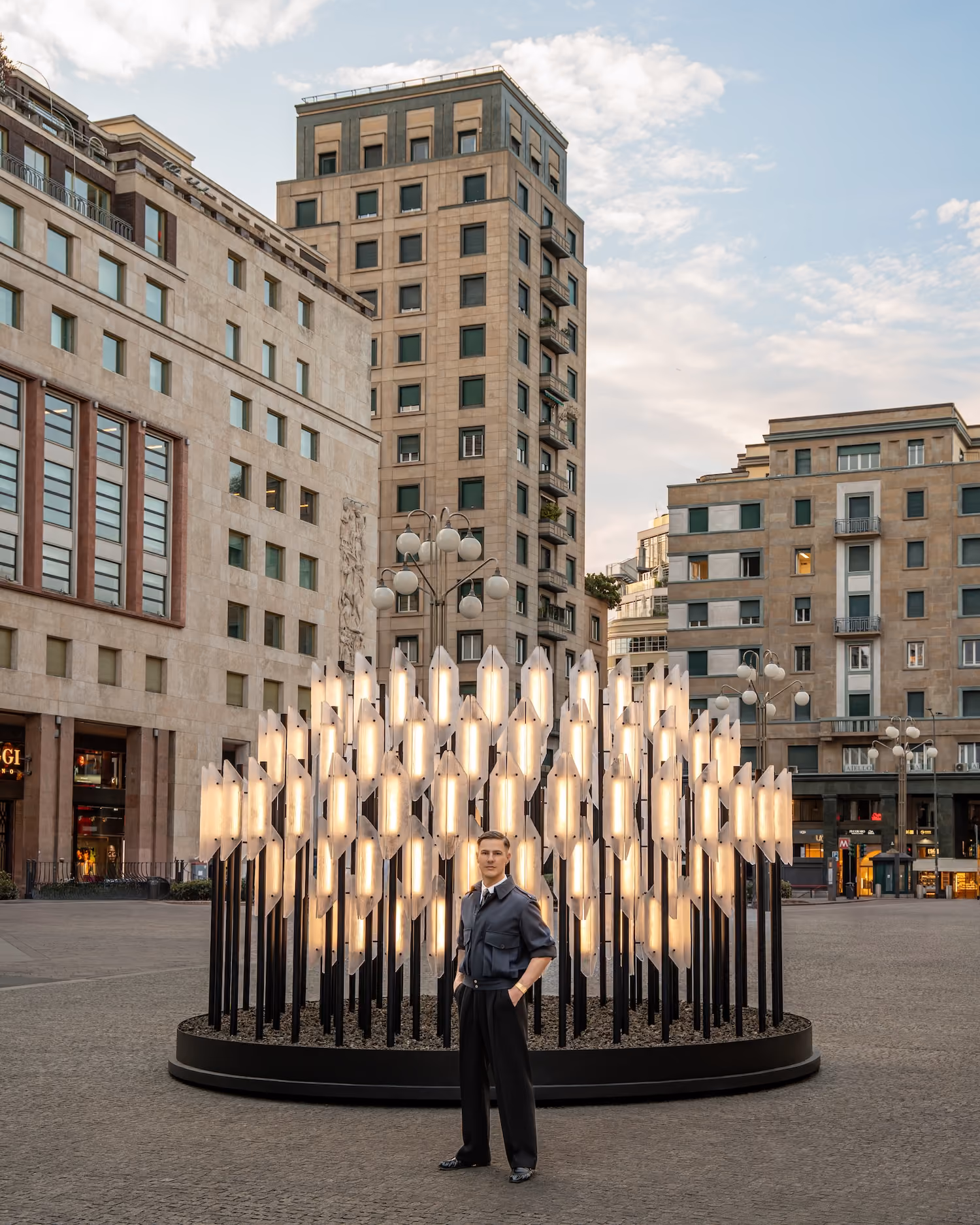 Man standing in front of a circular modern light installation in an urban plaza during sunset.