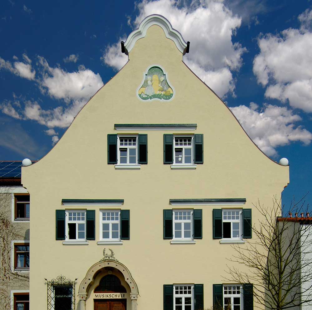 Fassade der Musikschule Pullach – ein gelbes Hauses mit geschwungenem Giebel, grünen Fensterläden und einem Wandbild über den Fenstern.