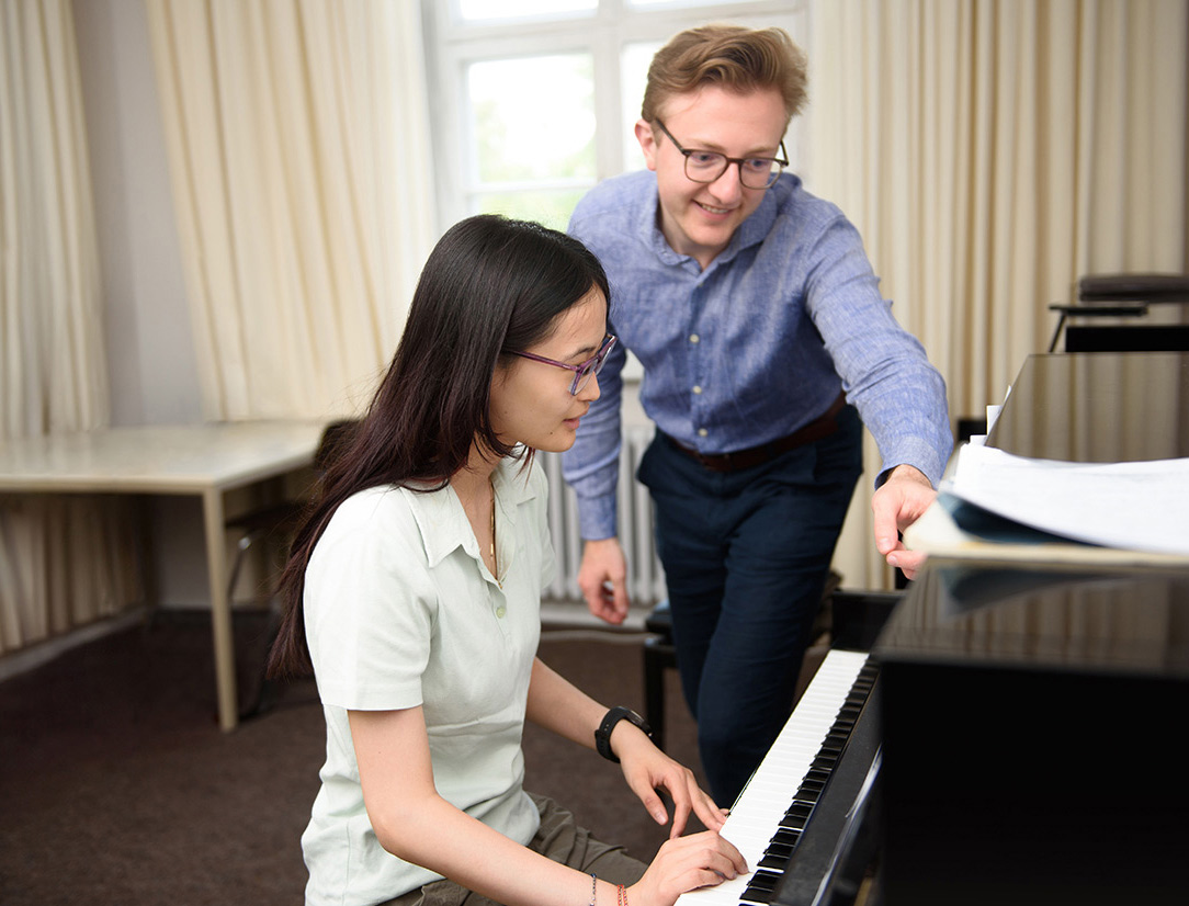 Ein Lehrer zeigt seiner Schülerin Noten am Klavier in einem hellen Raum.