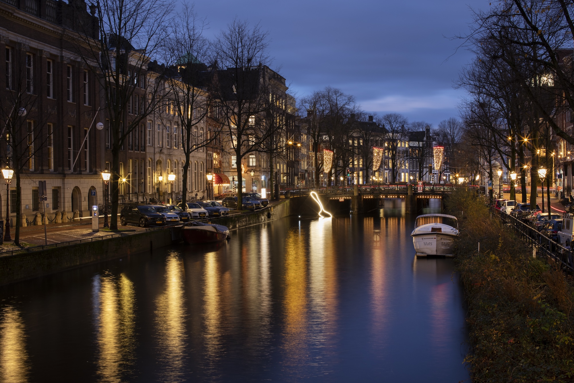 A canal at night shows lights reflecting off the surface and a light installation that is an outline of a spoon rests on the surface of the water.