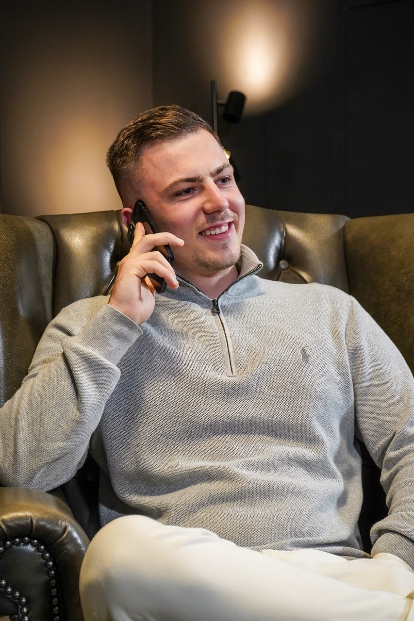 Junger Mann mit grauem Pullover sitzt in einem Sessel und telefoniert lächelnd.