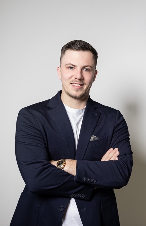 Junger Mann mit dunklem Sakko und weißem T-Shirt steht vor hellgrauem Hintergrund, lächelt und hat die Arme vor der Brust verschränkt.