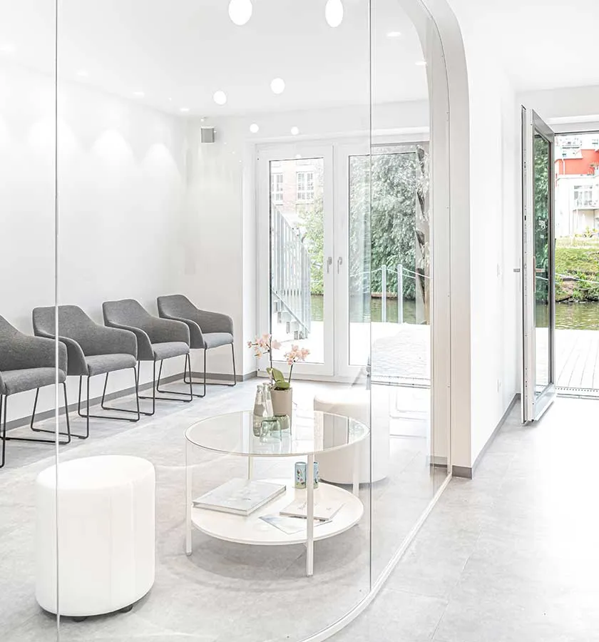 Heller Wartebereich mit grauen Stühlen, runden weißen Hockern und einem Glastisch mit Blumen und Magazinen vor großen Fenstern mit Blick auf Wasser.
