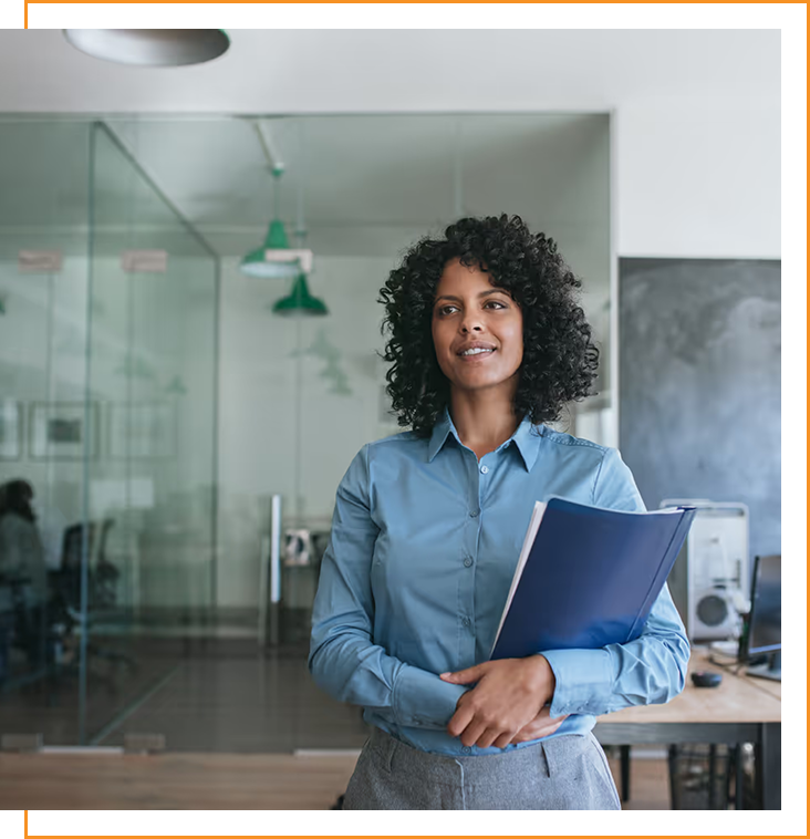 Confident woman in a blue shirt holding a blue folder standing in a modern office with glass walls.