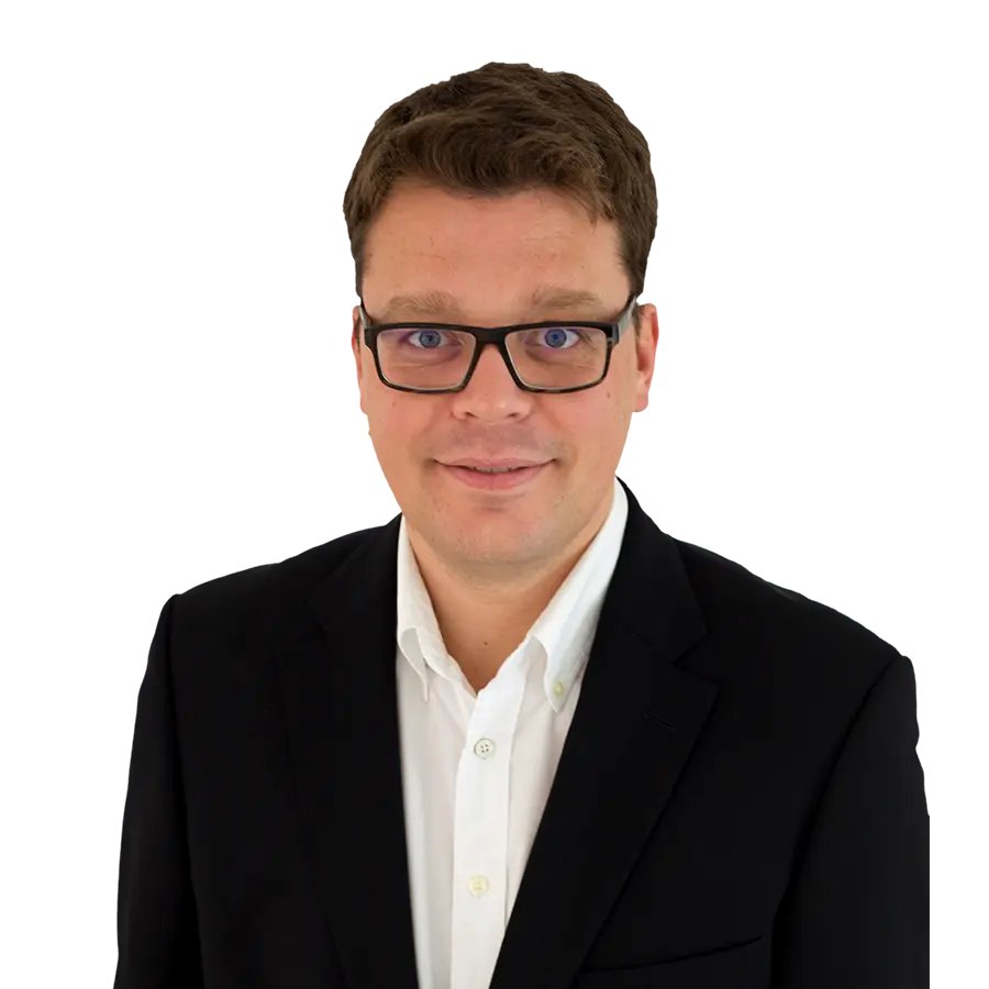Portrait von Sascha Bruchhaus im schwarzen Anzug und weißem Hemd, mit Brille und freundlichem Blick vor unscharfem Hintergrund – professionelles Businessfoto.