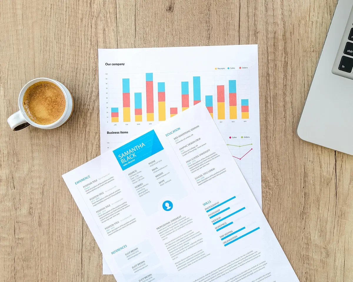 Auf einem Holztisch liegen ein Lebenslauf und ein Blatt mit bunten Balken- und Liniendiagrammen; daneben eine Tasse Kaffee und ein aufgeklappter Laptop – symbolisch für Bewerbung und Datenanalyse im Büroalltag.