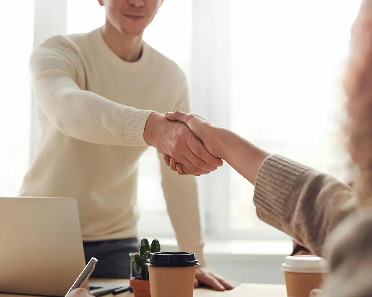 Zwei Personen geben sich zur Begrüßung oder zum Abschluss eines Gesprächs die Hand, im Vordergrund ein Kaffeebecher und ein Laptop – symbolisch für Vertrauen und professionellen Austausch.