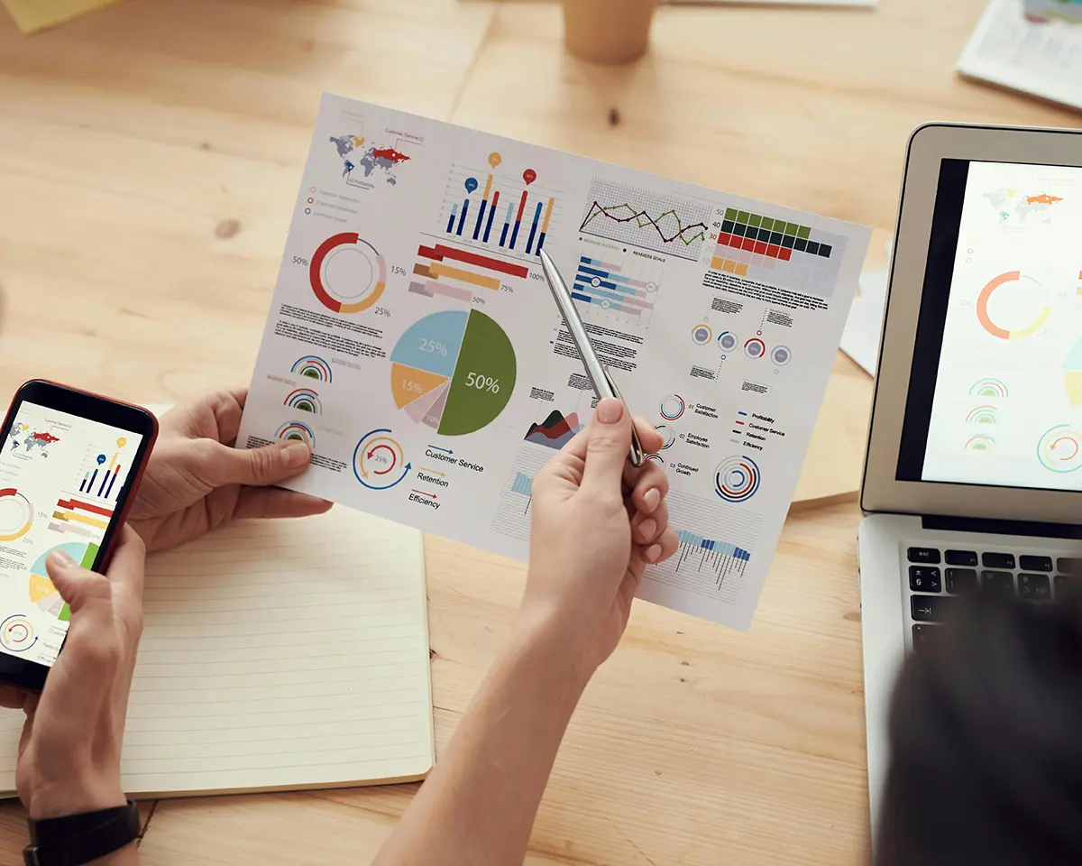 Zwei Personen analysieren bunte Diagramme auf einem Ausdruck, einem Smartphone und einem Laptopbildschirm – Fokus auf Datenvisualisierung und Zusammenarbeit