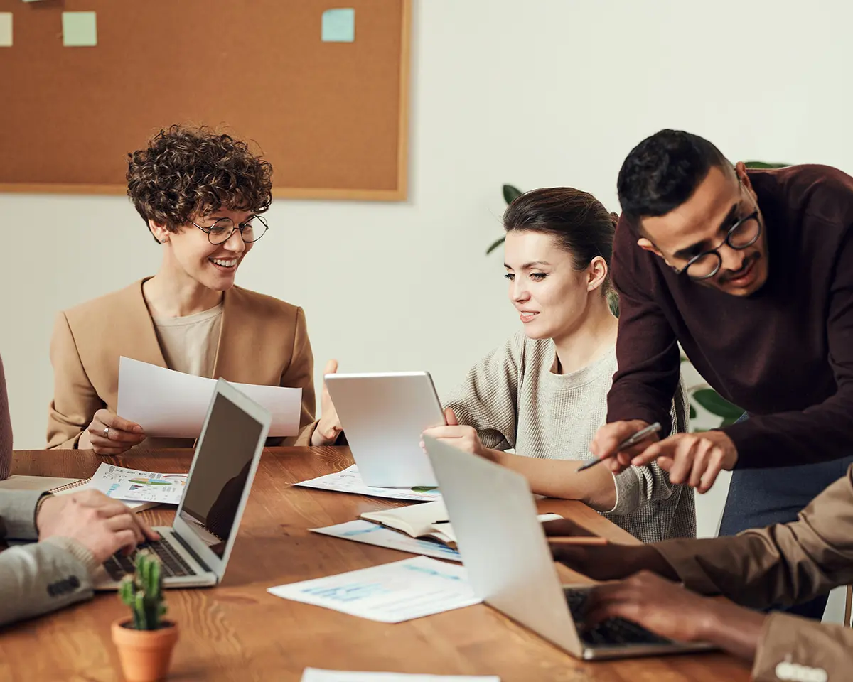 Ein Team aus mehreren Personen arbeitet gemeinsam an einem Holztisch; eine Frau hält ein Tablet, ein Kollege zeigt auf einen Laptopbildschirm, während eine weitere Person lächelnd in Unterlagen blickt.