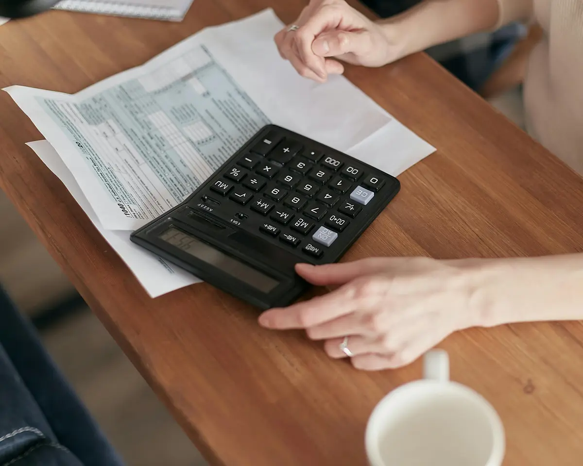 Person bedient einen Taschenrechner auf einem Holztisch, daneben liegen ein Formular mit Zahlen und eine weiße Kaffeetasse – typische Szene für Buchhaltung, Steuer oder Finanzplanung.