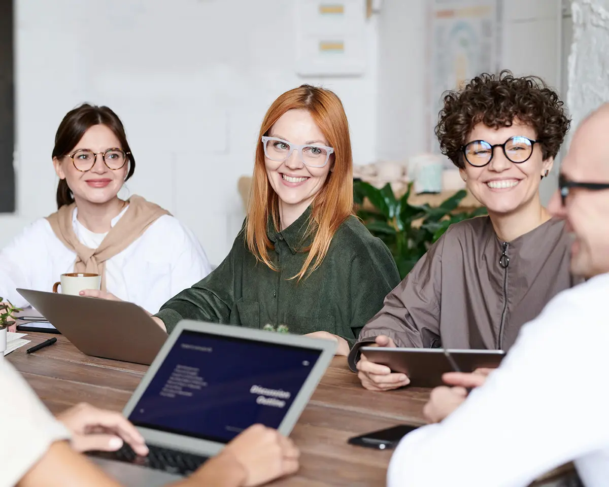 Drei Frauen sitzen lachend an einem Konferenztisch mit Laptops und Tablets, während eine weitere Person im Vordergrund mit ihnen spricht – freundliche Stimmung in einem kreativen Arbeitsumfeld.