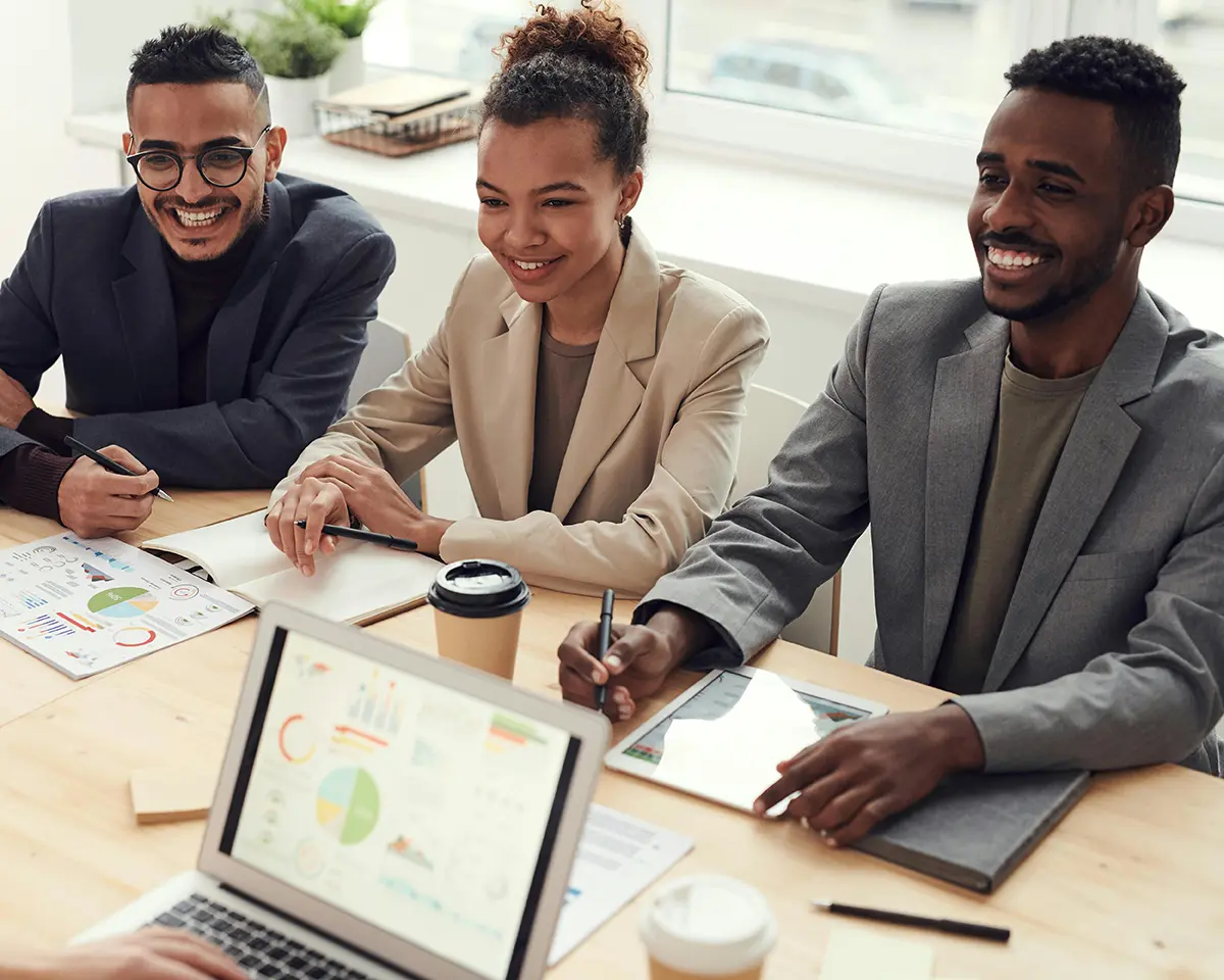 Drei Geschäftspersonen sitzen nebeneinander in einem Meetingraum, lächeln und machen sich Notizen; auf dem Tisch liegen Diagramme, Laptops und Kaffeebecher