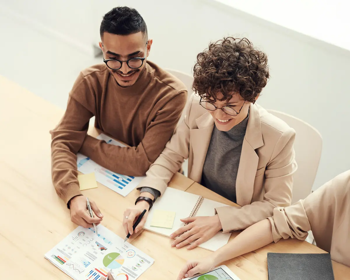Zwei junge Personen sitzen nebeneinander an einem Holztisch und analysieren gemeinsam bunte Diagramme auf einem Papier – lockere und konzentrierte Arbeitsatmosphäre im Team.