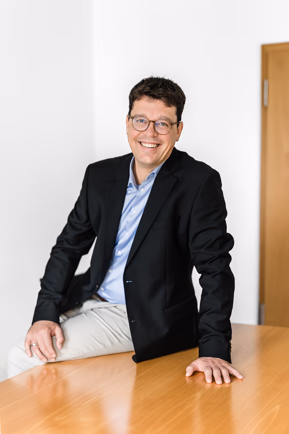 Smiling man wearing glasses and a black blazer sitting on a wooden table in a bright room.