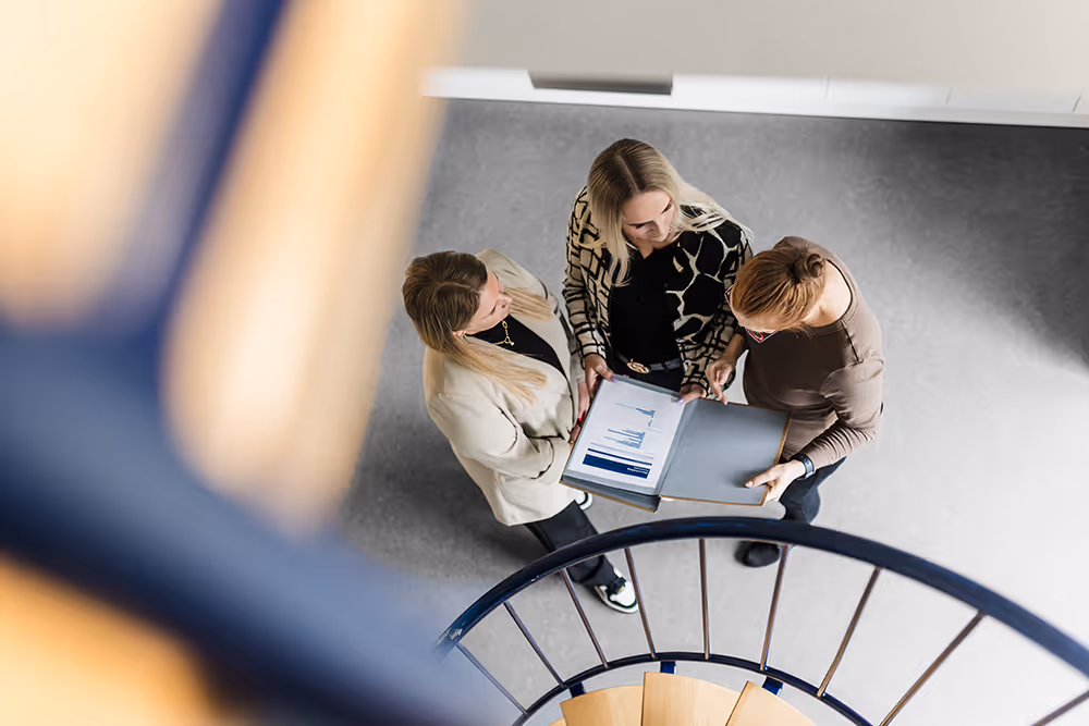 Three women standing by a spiral staircase, looking at documents in a folder.