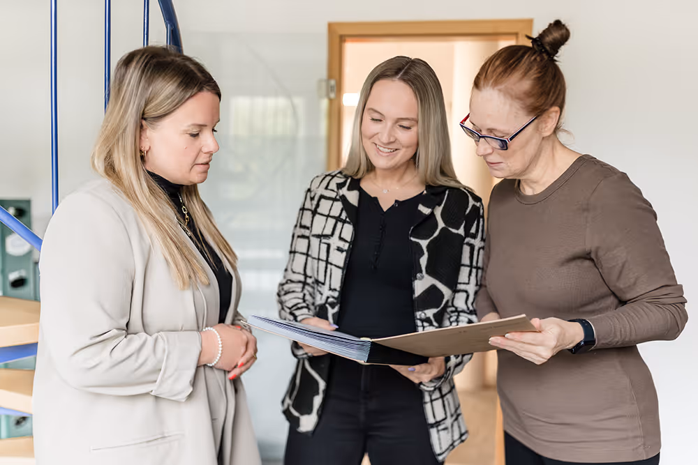 Three women standing indoors, looking at and discussing content in an open folder.
