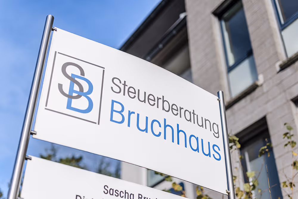White signboard with gray and blue logo and text reading 'Steuerberatung Bruchhaus' against blue sky and building background.