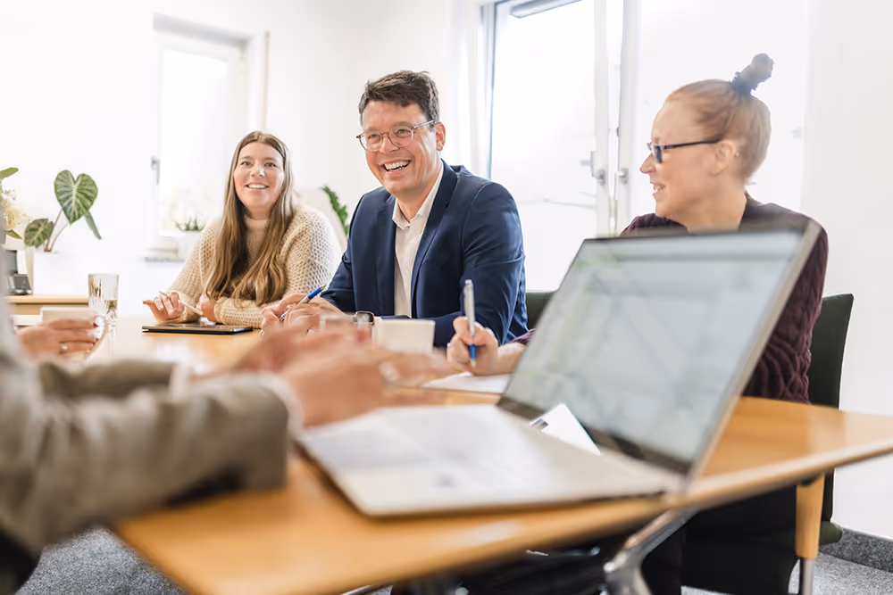 Three people smiling and engaged in a meeting around a table with a laptop in the foreground.