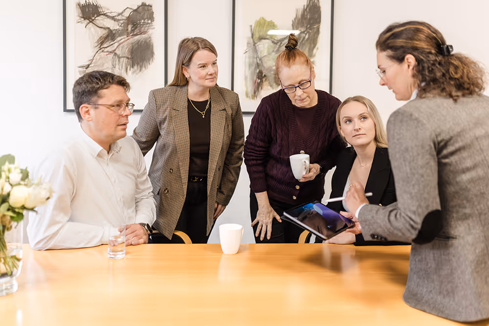 Five people engaged in a discussion around a wooden table with two white mugs and a glass of water, one person holding a digital tablet with a stylus.
