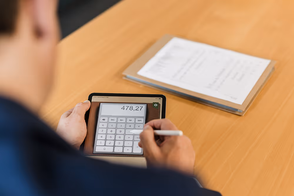 Person using a stylus to operate a calculator app on a tablet, with a stack of documents on a wooden table.