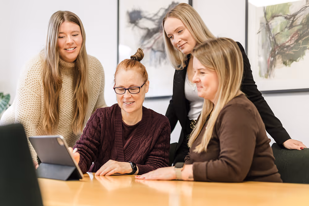 Four women gathered around a tablet on a wooden table, engaged in a discussion in an office setting.