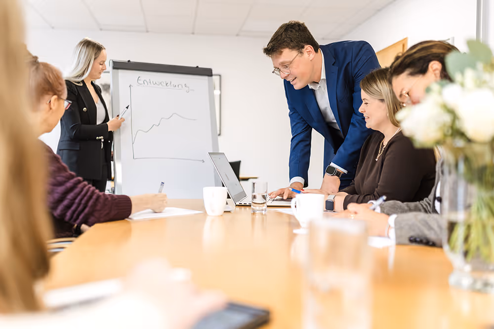 A group of people in a meeting room with a woman presenting a rising line chart on a flip chart, while others take notes and interact around a table.