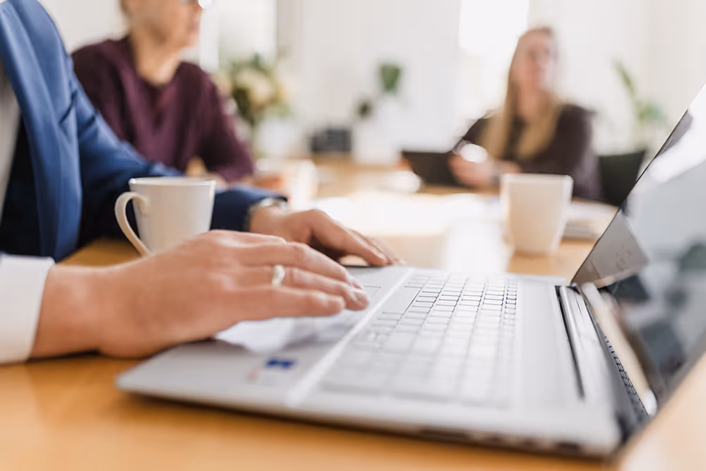 Person typing on a laptop keyboard at a table with blurred people and coffee cups in the background.