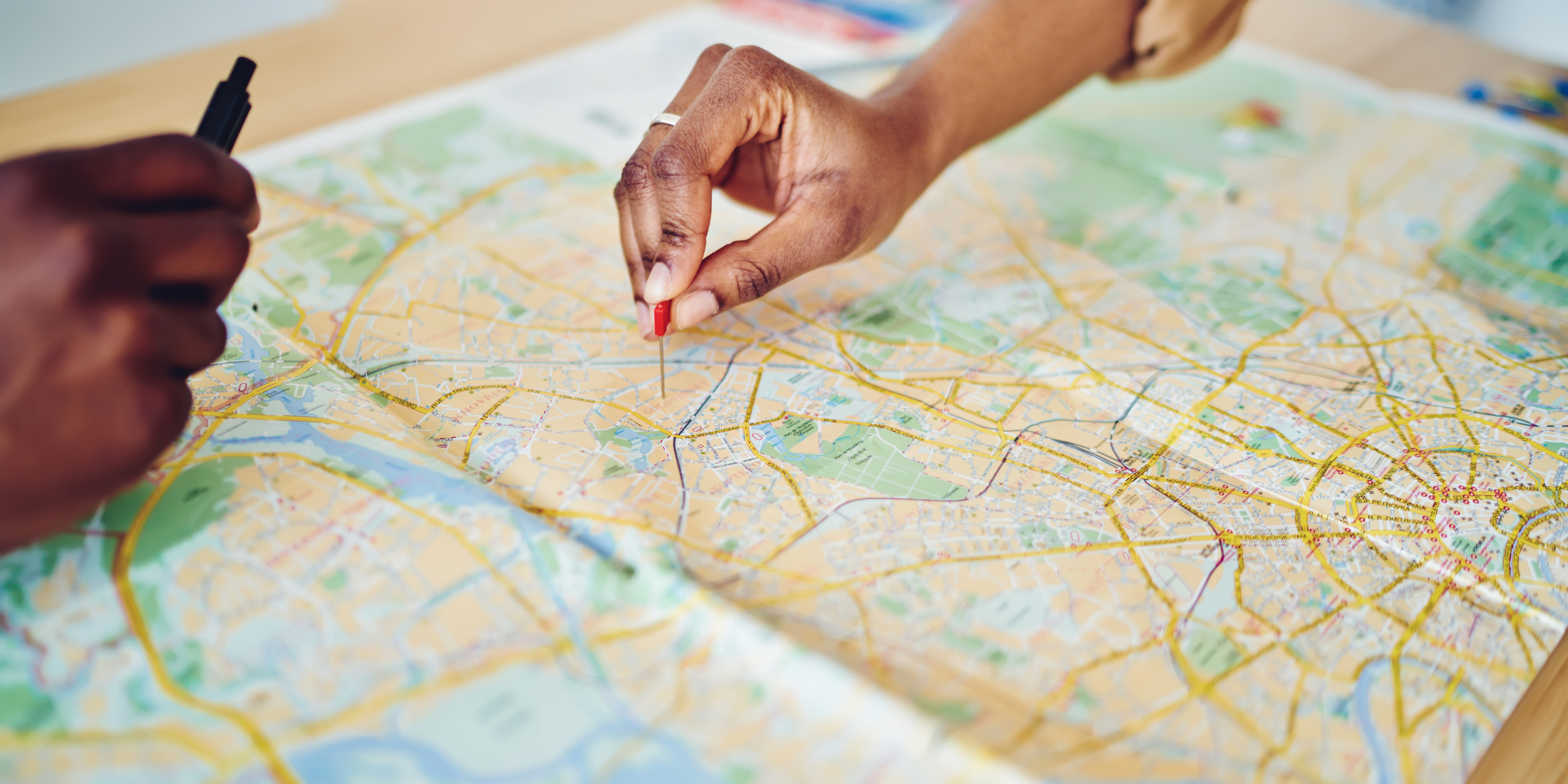 Paper map with one hand placing a red push pin and another and holding a pencil