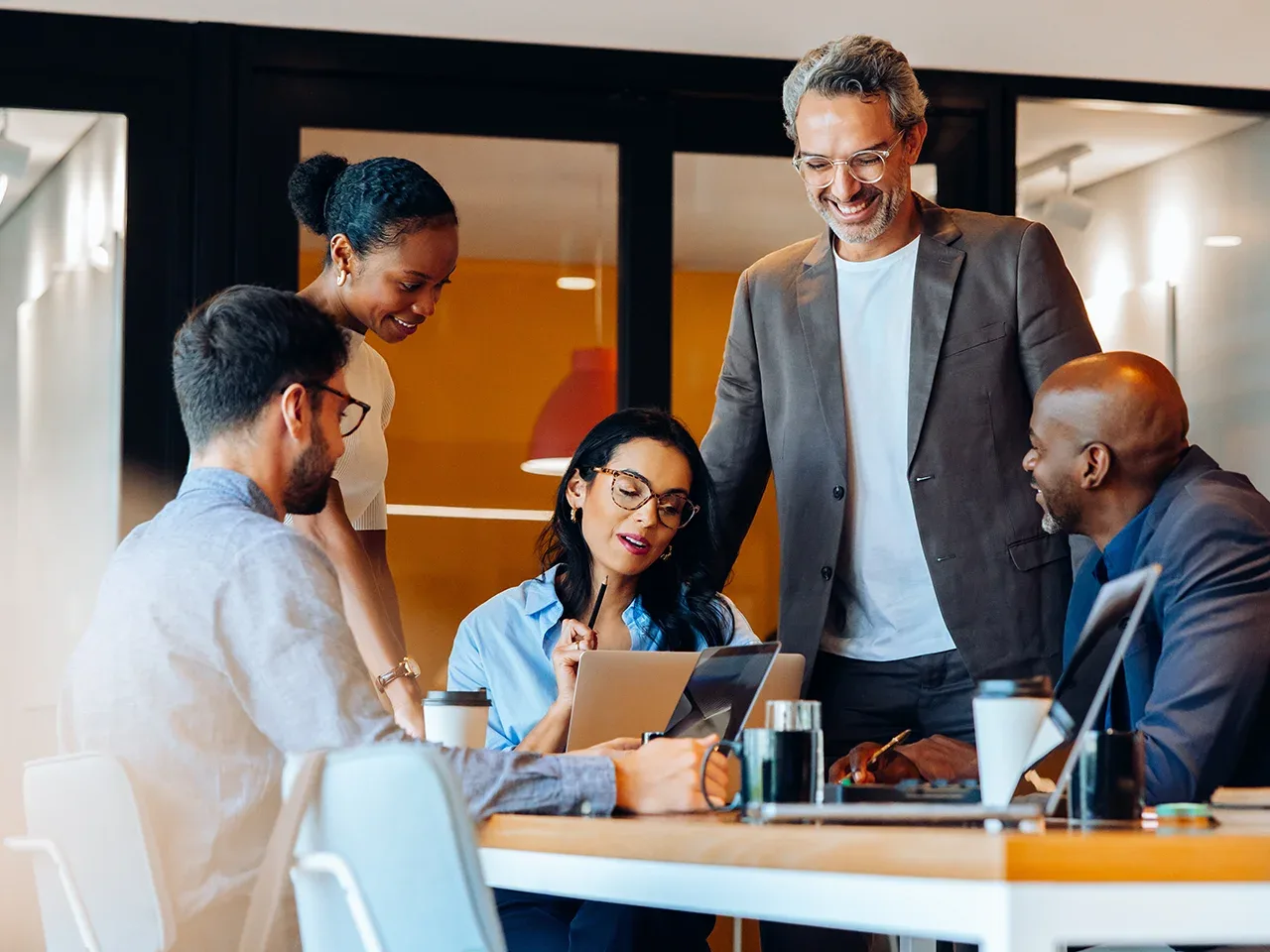 Five diverse colleagues collaborating around a table with laptops and coffee cups in a modern office.