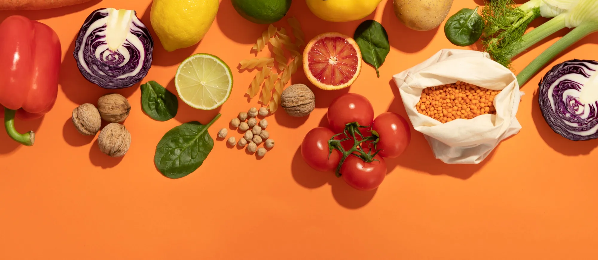 Assortment of fresh vegetables, fruits, nuts, and legumes including red bell pepper, red cabbage halves, lemons, lime slices, tomatoes, walnuts, chickpeas, red lentils in a cloth bag, fusilli pasta, spinach leaves, and fennel on an orange background.