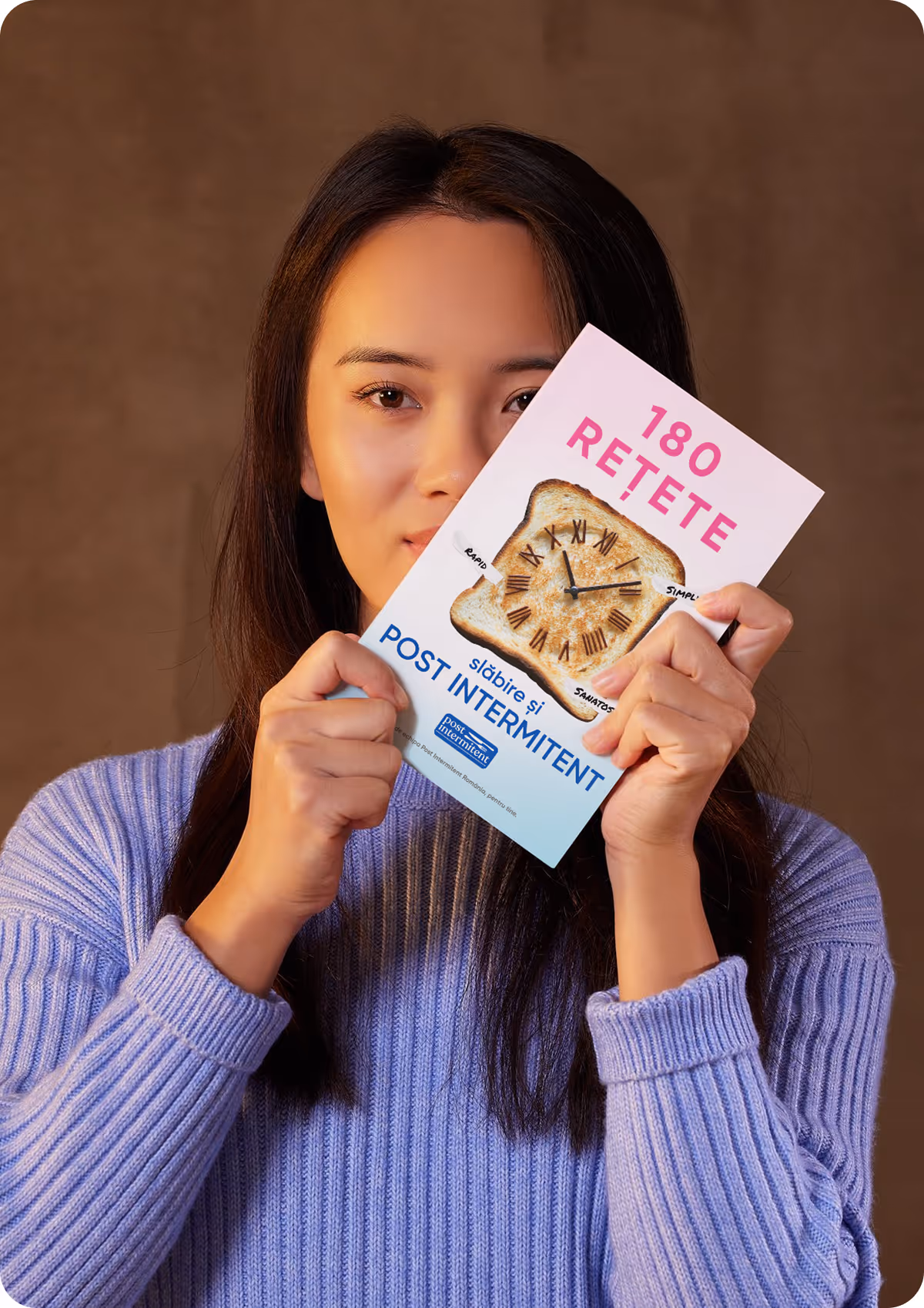 O femeie cu pulover mov ține în fața feței o carte intitulată „180 rețete slăbire și post intermittent” cu o felie de pâine prăjită ilustrată ca un ceas.