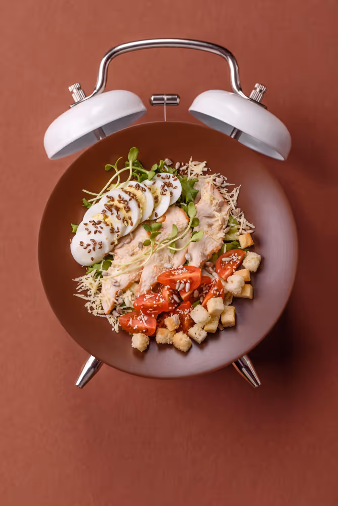 Salată cu felii de ou fiert, piept de pui, roșii cherry, crutoane, semințe și salată verde, într-o farfurie în formă de ceas cu alarmă albă și maro.