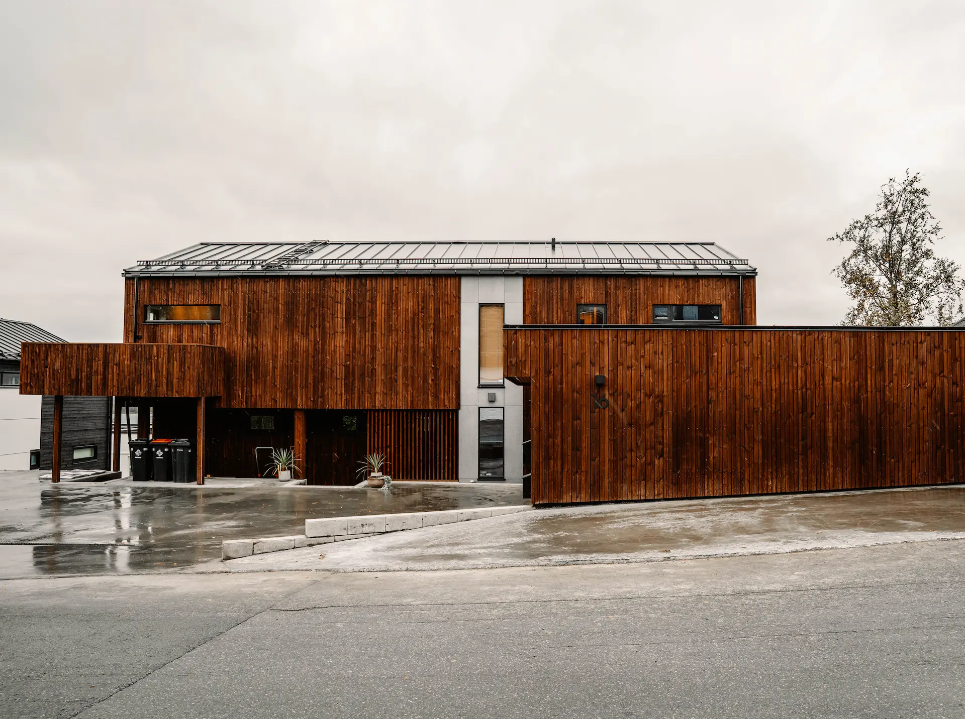 Nybygd bolig med mørk trekledning og carport, fotografert på en våt høstdag