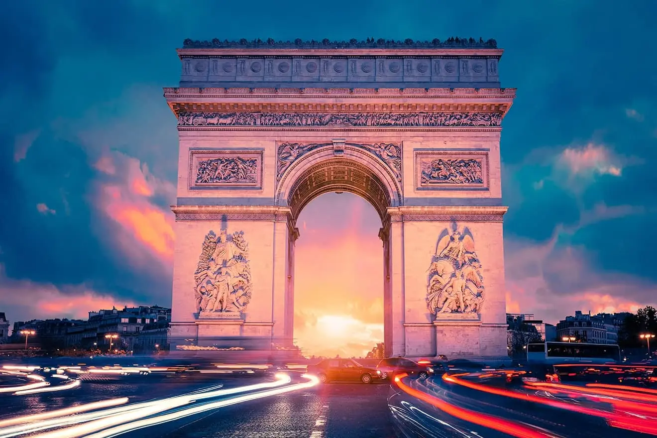 The Arc de Triomphe in Paris at sunset, with the sky glowing in shades of blue and pink. In the foreground, the lights of passing cars leave long, dynamic trails of light across the roundabout.